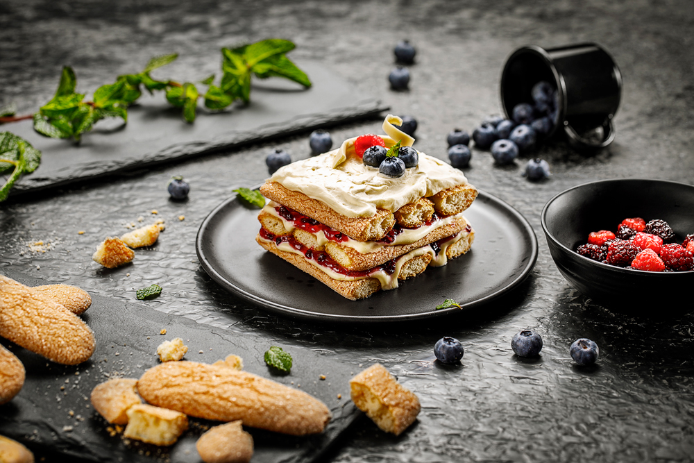 Biscoitos champanhe dourados empilhados em prato preto, cobertos com nata e frutas vermelhas, sobre mesa escura.