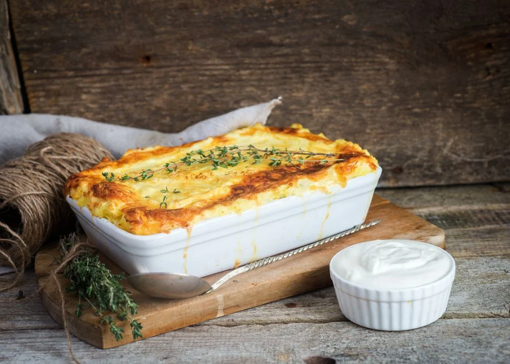Travessa branca com batatas gratinadas, cobertas por queijo derretido, servida sobre mesa de madeira.