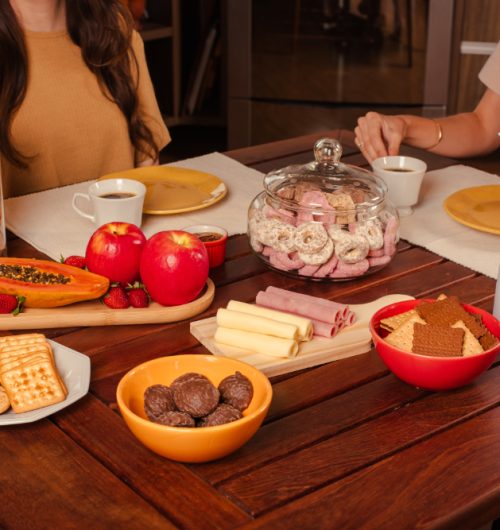 Mesa de café da manhã com xícara de chá, pratos, queijo, presunto, rosquinhas e biscoitos, acompanhados de frutas.