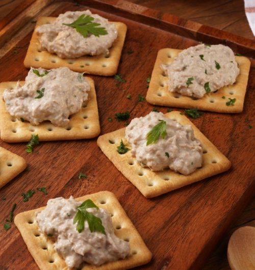Biscoitos cream cracker sobre tábua de madeira com porções de patê e salsinha.