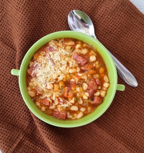 Tigela com canjica salgada com calabresa, queijo ralado e ervas, servida sobre mesa marrom.