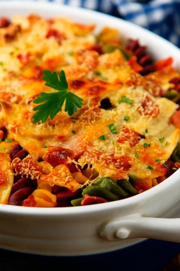 Prato de macarrão sabor vegetal gratinado com frango e coberto por queijo dourado, servido em travessa branca sobre fundo desfocado.