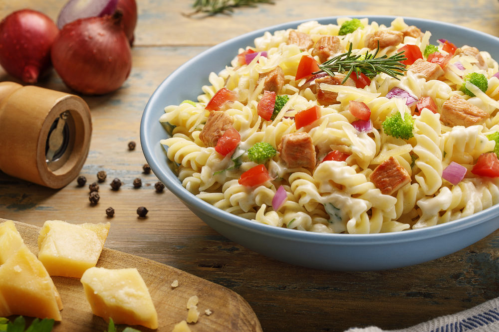 Prato fundo com macarrão parafuso com frango, cebola roxa, tomates, brócolis e queijo, sobre mesa de clara.