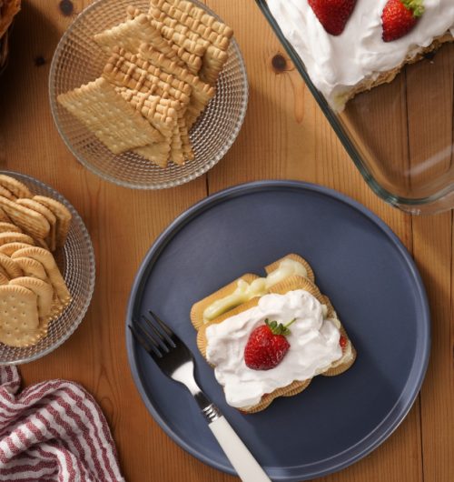 Mesa composta por biscoitos maisena, acompanhados de copo de leite, geleia, pãos tostados e sobremesa de morango, dispostos sobre mesa de madeira.