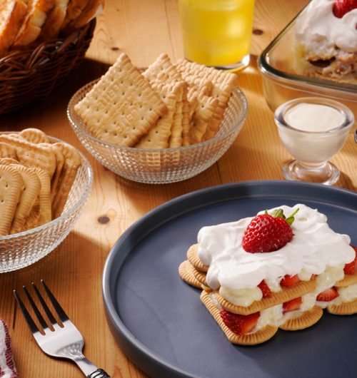 Mesa composta por biscoitos maisena, acompanhados de copo de leite, geleia, pãos tostados e sobremesa de morango, dispostos sobre mesa de madeira.