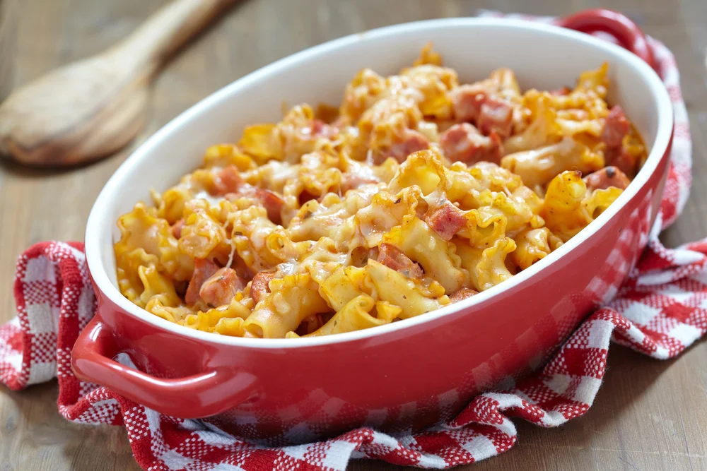Travessa vermelha com macarrão espiral gratinado ao forno, coberto com bacon e mussarela derretida, servida sobre mesa de madeira.
