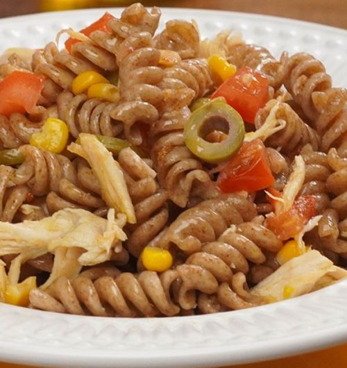 Prato de macarrão parafuso com frango, tomates, azeitonas e milhos, servido em prato branco sobre fundo neutro.