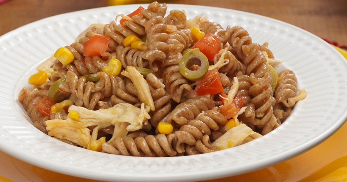 Prato de macarrão parafuso com frango, tomates, azeitonas e milhos, servido em prato branco sobre fundo neutro.