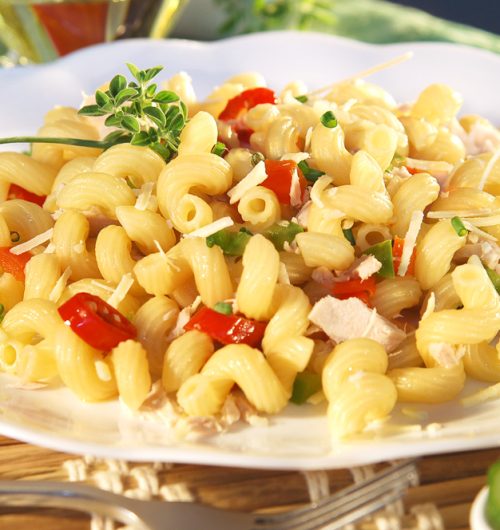 Prato de macarrão espiral com frango, tomate e pimentão verde, coberto com queijo e cebolinha, servido em prato branco; fundo desfocado.