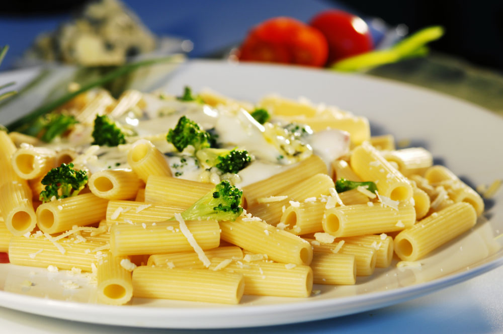 Prato de macarrão rigatoni, coberto com molho branco, brócolis e queijo ralado, disposto em um prato branco e fundo desfocado.