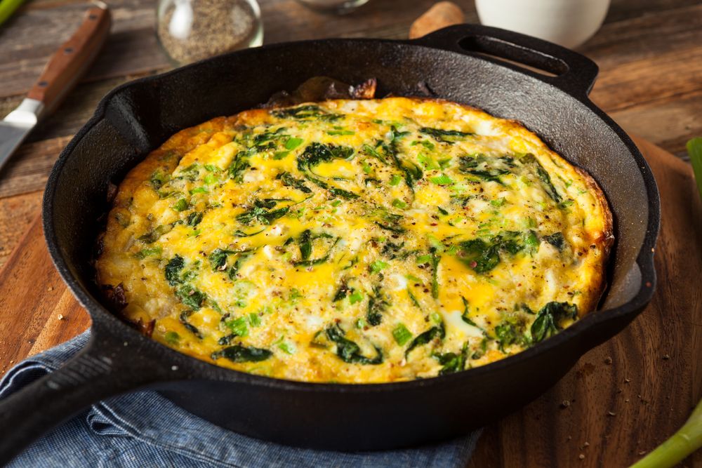 Prato de omelete com espinafre e queijo ralado, servido em frigideira sobre mesa de madeira.