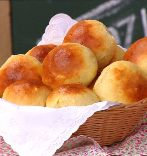 Cesta de pãozinhos de batata doce dourados, disposta sobre mesa de madeira.