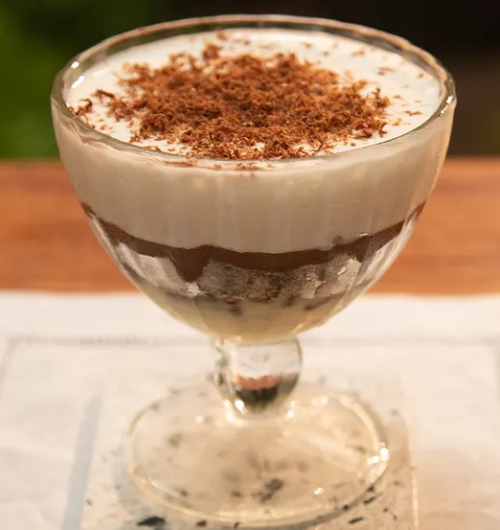 Taça de pavê de chocolate com wafer e creme, coberto por raspas de chocolate, sobre mesa de madeira.