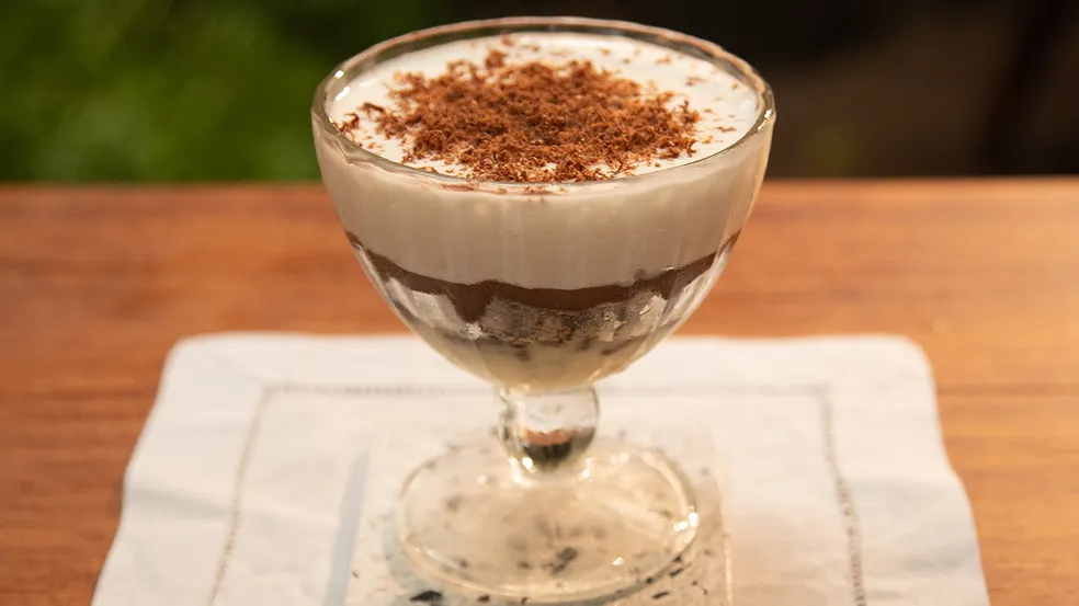 Taça de pavê de chocolate com wafer e creme, coberto por raspas de chocolate, sobre mesa de madeira.