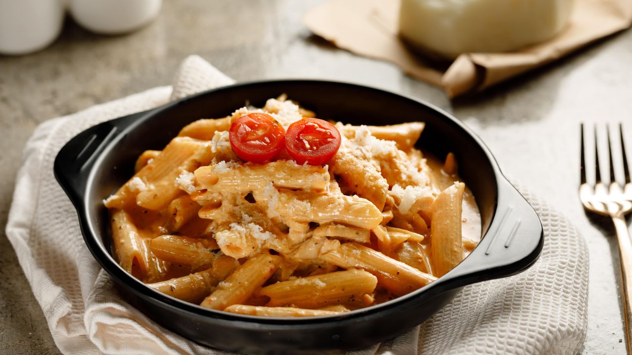 Prato fundo preto com macarrão penne ao molho cremoso, servido com queijo ralado e tomates, sobre mesa de madeira clara.