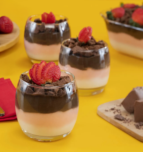 Taça de vidro transparente com sobremesa cremosa de rosquinha, decorada com morangos e calda de chocolate, sobre fundo amarelo.