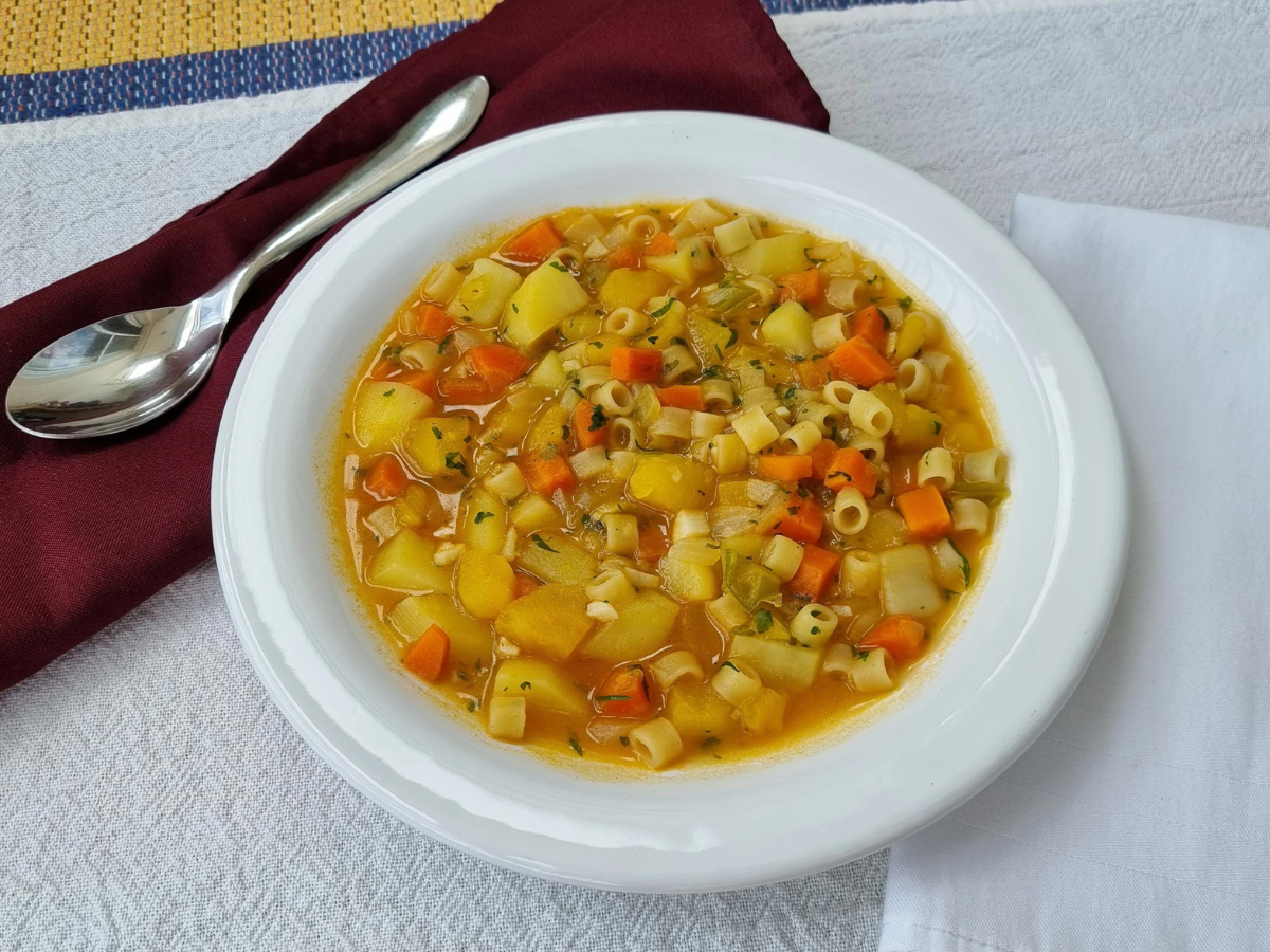 Tigela com sopa de legumes contendo massa ave-maria, cenoura, batata e vagem em caldo, servida sobre mesa clara.