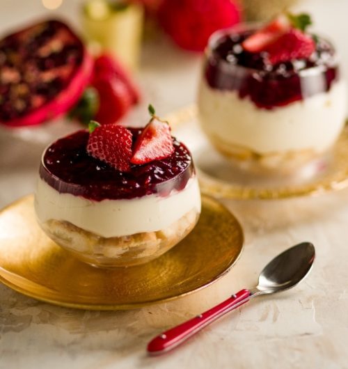 Taça de vidro transparente com camadas de chantilly, frutas vermelhas e biscoito champanhe, com morangos em cima, sobre mesa clara.
