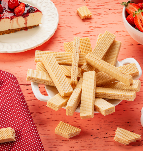 Biscoitos Wafer sabor morango, ao lado da fatia de cheesecake de morango, sobre mesa rosa.