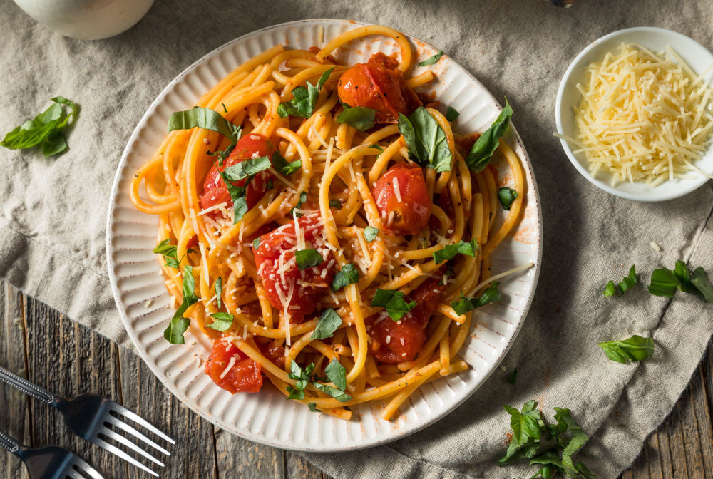 Prato de espaguete coberto com tomates, manjericão e queijo, disposto em prato branco sobre mesa clara.