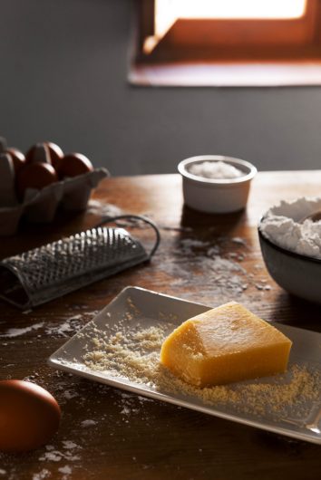 Queijo em pedaço triangular sobre mesa de madeira, ao lado de ralador metálico com porção de queijo ralado acumulada na base.