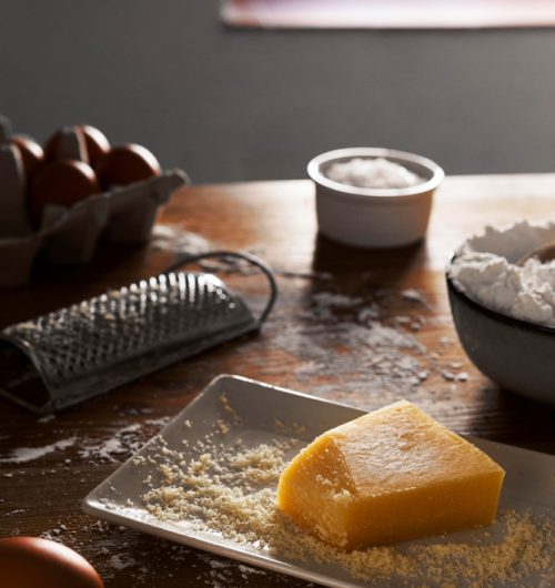 Queijo em pedaço triangular sobre mesa de madeira, ao lado de ralador metálico com porção de queijo ralado acumulada na base.