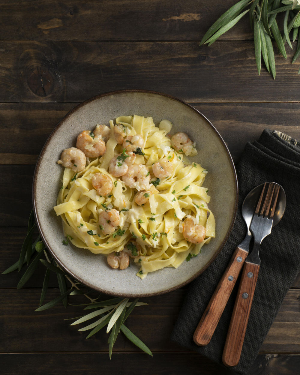Prato de macarrão fettuccine servido com camarão, decorado com manjericão, em prato branco sobre mesa de madeira.