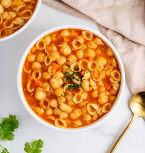 Sopa servida em prato fundo branco com macarrão tipo concha, legumes e caldo, sobre mesa clara.