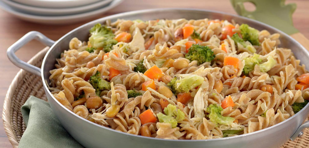 Prato de macarrão integral com pedaços de frango, cenoura, brócolis e feijão, servido em panela clara sobre fundo desfocado.