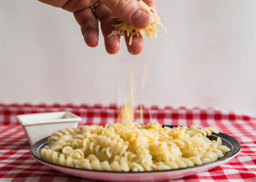 Mão polvilhando queijo ralado sobre prato de macarrão, servido em prato branco sobre toalha xadrez.