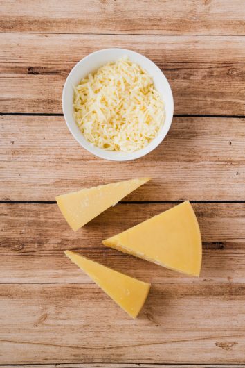 Queijo triangular em peça grande ao lado de porção de queijo ralado, dispostos sobre mesa de madeira.