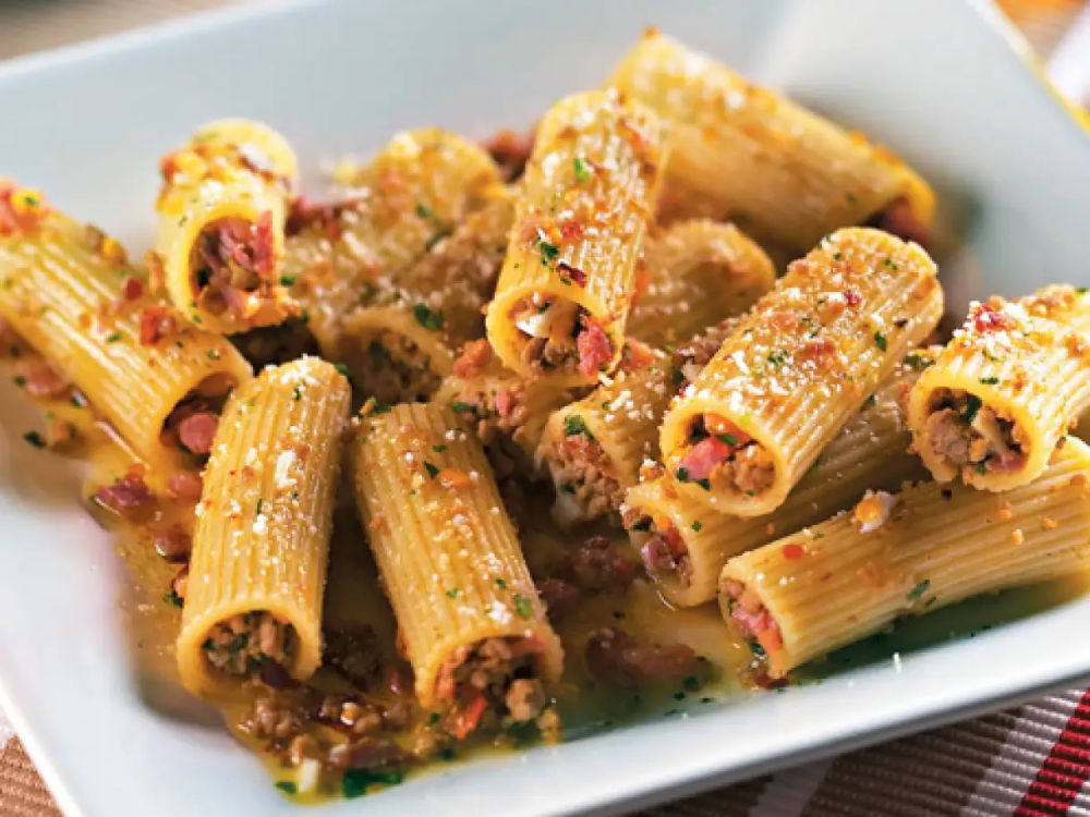 Prato de macarrão rigatoni servido em prato branco, coberto com carne moída e bacon, decorado com manjericão fresco.